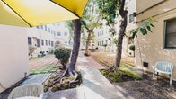 This property is defined by its expansive central courtyard, which serves as the heart of the space. The outdoor area is framed by light-colored stucco building facades on multiple sides, creating an enclosed yet open atmosphere that invites natural light throughout the day. Mature trees with prominent surface roots are planted throughout the courtyard, providing natural shade and a sense of established greenery that softens the hardscape surroundings. The ground is finished with a combination of brick-toned pavers and concrete slabs, forming organized walkways and gathering zones. Multiple outdoor seating arrangements are distributed across the courtyard, each shaded by patio umbrellas in yellow, navy, and white, making the space well-suited for casual gatherings or relaxed outdoor use. Potted plants and trimmed shrubs are positioned along the building perimeters, adding further greenery and texture. An arched doorway detail is visible on one of the building wings, offering a subtle architectural accent. Bicycle parking is tucked along a side passage between structures, suggesting a practical, community-oriented environment. Overall, the courtyard presents a functional and welcoming outdoor setting with a relaxed, urban character.