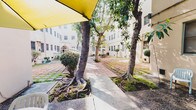 This property is defined by its expansive central courtyard, which serves as the heart of the space. The outdoor area is framed by light-colored stucco building facades on multiple sides, creating an enclosed yet open atmosphere that invites natural light throughout the day. Mature trees with prominent surface roots are planted throughout the courtyard, providing natural shade and a sense of established greenery that softens the hardscape surroundings. The ground is finished with a combination of brick-toned pavers and concrete slabs, forming organized walkways and gathering zones. Multiple outdoor seating arrangements are distributed across the courtyard, each shaded by patio umbrellas in yellow, navy, and white, making the space well-suited for casual gatherings or relaxed outdoor use. Potted plants and trimmed shrubs are positioned along the building perimeters, adding further greenery and texture. An arched doorway detail is visible on one of the building wings, offering a subtle architectural accent. Bicycle parking is tucked along a side passage between structures, suggesting a practical, community-oriented environment. Overall, the courtyard presents a functional and welcoming outdoor setting with a relaxed, urban character.