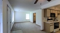 This single-floor space offers a straightforward and functional layout designed with practicality in mind. The main room is generously proportioned, featuring wall-to-wall carpeting that lends a warm, comfortable feel underfoot. Bright white walls and a clean, smooth ceiling create a light and airy atmosphere, while natural light filters in through multiple windows, enhancing the sense of openness. A ceiling fan is centrally positioned overhead, providing both circulation and a subtle decorative element.

A doorway off the main room leads into a dedicated walk-in closet, offering a substantial amount of organized storage space — a highly practical feature that adds to the overall functionality of the layout. To one side of the room, a built-in cabinetry section with warm wood-toned finishes provides additional storage and counter surface, serving as a versatile utility area.

The overall ambiance of the space is clean, neutral, and move-in ready, with a minimalist aesthetic that provides an excellent blank canvas for personalization. The combination of natural light, generous room dimensions, ample storage, and thoughtful built-in features makes this a highly practical and adaptable single-floor space.