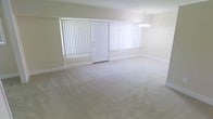 This single-story space features an open and airy living area designed with simplicity and functionality in mind. The room is finished with wall-to-wall neutral-toned carpeting that lends a clean, understated aesthetic throughout. Light-colored walls complement the soft flooring, creating a cohesive and calm environment.

Natural light fills the space through two large windows dressed with vertical blinds, allowing for flexible light control. A central white paneled door provides access to the exterior, flanked by these generous windows that enhance the sense of openness. An elegant multi-bulb chandelier is positioned toward one end of the room, serving as a refined focal point and providing ambient overhead lighting.

The layout offers a notably open floor plan, providing ample space for a variety of furniture arrangements and uses. Wide baseboards and clean wall finishes contribute to the room's polished, well-maintained appearance. The overall ambiance is bright, neutral, and move-in ready, offering a versatile canvas suitable for a range of interior design styles.