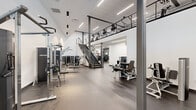 Located at 2939 Main St in Buffalo, NY, this contemporary fitness facility offers a well-appointed, multi-level training environment designed for a comprehensive workout experience. The main floor is expansive and open, bathed in natural light through floor-to-ceiling black-framed windows along the exterior wall, and features polished concrete-style flooring throughout. The soaring ceilings on the main level create an airy, energizing atmosphere, with sleek pendant and linear LED lighting complementing the clean white walls and steel structural columns.

The main training floor is outfitted with a wide array of strength machines, cable systems, free weights, and benches, providing ample space for both machine-based and functional training. A dedicated functional training room offers mirrored walls, organized storage racks stocked with kettlebells, medicine balls, resistance bands, BOSU balls, and suspension trainers — ideal for dynamic, varied workouts.

A steel staircase with cable railings leads to the mezzanine level, where a row of cardio machines, including ellipticals and steppers, is neatly arranged beneath a drop ceiling with linear LED lighting. The industrial-modern design aesthetic is consistent throughout, with a neutral palette of white, grey, and black creating a focused, professional training environment.