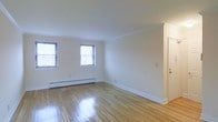 This single-floor space presents a clean, minimalist aesthetic that is both inviting and functional. The main living area is highlighted by polished hardwood floors in a warm honey tone, which reflect natural light and lend the room a bright, open feel. White walls and white trim throughout maintain a crisp, neutral palette, making the space easy to personalize. Crown molding lines the ceiling, adding a subtle architectural detail that elevates the overall character of the room. Two windows along the exterior wall allow ample natural light to filter in, enhancing the airy atmosphere. A baseboard heating system runs beneath the windows, ensuring comfortable warmth throughout the colder months. The smooth, white ceiling and clean corners contribute to a well-maintained and move-in-ready condition. A doorway visible along one wall hints at additional rooms or storage beyond, suggesting a practical and connected layout. Overall, the space combines classic details — from the hardwood flooring to the crown molding — with a straightforward, light-filled design that provides a comfortable and adaptable living environment.