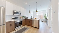 This thoughtfully designed one-bedroom, one-bathroom residence offers a cohesive modern aesthetic across its single floor. Light wide-plank hardwood flooring flows continuously throughout, tying together each room seamlessly.

The open-concept kitchen is a standout feature, outfitted with two-tone cabinetry — crisp white upper cabinets paired with warm wood-grain lowers — and a full suite of stainless steel appliances. A peninsula with a white quartz countertop provides bar seating and separates the kitchen from the living area. Sleek black conical pendant lights add a contemporary focal point overhead.

The living room benefits from large windows that invite generous natural light and offer views of the surrounding urban streetscape. Neutral walls and light flooring create a bright, airy atmosphere throughout.

The bedroom is a calm retreat, featuring additional large windows, a built-in closet with shelving, and continued light hardwood floors. Its understated palette ensures a restful environment.

The bathroom is finished to a high standard, with full marble-look tile covering the walls and floors, a floating wood-grain vanity, a soaking tub, and matte black fixtures and hardware that lend a refined, modern contrast to the bright surroundings.