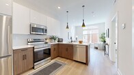 This thoughtfully designed one-bedroom, one-bathroom residence offers a cohesive modern aesthetic across its single floor. Light wide-plank hardwood flooring flows continuously throughout, tying together each room seamlessly.

The open-concept kitchen is a standout feature, outfitted with two-tone cabinetry — crisp white upper cabinets paired with warm wood-grain lowers — and a full suite of stainless steel appliances. A peninsula with a white quartz countertop provides bar seating and separates the kitchen from the living area. Sleek black conical pendant lights add a contemporary focal point overhead.

The living room benefits from large windows that invite generous natural light and offer views of the surrounding urban streetscape. Neutral walls and light flooring create a bright, airy atmosphere throughout.

The bedroom is a calm retreat, featuring additional large windows, a built-in closet with shelving, and continued light hardwood floors. Its understated palette ensures a restful environment.

The bathroom is finished to a high standard, with full marble-look tile covering the walls and floors, a floating wood-grain vanity, a soaking tub, and matte black fixtures and hardware that lend a refined, modern contrast to the bright surroundings.