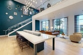 Located at 925 W 20th St in Houston's vibrant Heights neighborhood, this thoughtfully designed two-story communal space offers a rich blend of modern architecture and curated interior design. The expansive main floor is anchored by a striking large-format island with a white quartz top and dark base, surrounded by bar-height seating beneath a cascading cluster of globe pendant lights that draw the eye upward toward soaring double-height ceilings. Bold teal accent walls adorned with eclectic framed artwork and decorative mirrors add depth and personality throughout. A glass-railed staircase leads to the upper mezzanine level, which features built-in bar seating, additional lounge chairs, and gallery-style wall art, all overlooking the main floor below. Floor-to-ceiling windows along the exterior wall flood the lounge area with abundant natural light and offer views of the outdoor greenery. The main floor also includes a cozy lounge with tufted leather sofas, a charcoal-walled banquette nook with ornate framed prints, and a dedicated workspace with built-in desks and storage finished in teal cabinetry. Light wood flooring flows throughout both levels, unifying the space with warmth and continuity. This multi-functional environment balances social gathering, focused work, and relaxed lounging in one cohesive, design-forward setting.