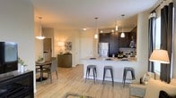This single-floor residence offers a thoughtfully designed open-concept layout that seamlessly connects the kitchen, dining, and living areas into one cohesive and functional living space. The kitchen is a standout feature, equipped with dark espresso cabinetry, stainless steel appliances including a full-size refrigerator and microwave, and a generous granite-topped island with bar seating for multiple guests. Pendant lighting suspended above the island adds a refined, modern touch. The dining area sits adjacent to the kitchen, providing an intimate setting with room for a full dining table and chairs. Warm, light wood-style flooring flows consistently throughout, creating a welcoming and unified aesthetic. Neutral-toned walls enhance the brightness of the space and complement the contemporary design elements. Track lighting and decorative pendant fixtures ensure the entire area is well-lit and inviting. A hallway visible beyond the dining area suggests access to additional private rooms, maintaining a clear separation between the social and personal zones of the residence. Overall, this space combines modern finishes with a practical, open layout ideal for both everyday living and entertaining.