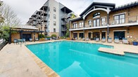 Located at 245 E Broad St in the heart of downtown Greenville, SC, this outdoor amenity space offers a resort-style pool experience designed for relaxation and recreation. The centerpiece is a generously sized swimming pool with crystal-clear blue water, featuring a shallow entry point and a built-in step section that creates a multi-depth layout ideal for a range of users. The pool deck is finished in a light-toned concrete surround, providing ample space for lounging with multiple chaise lounge chairs and seating arrangements positioned throughout. Two dark-framed pergolas anchor distinct zones on the deck, offering shaded retreats for outdoor dining and casual gatherings. A dedicated grilling area equipped with gas grills and café-style table-and-chair sets makes the space well-suited for social entertaining. Lush mature trees and manicured shrubs line the perimeter, creating a sense of privacy and a natural green backdrop. The space is framed by the building's classic brick and contemporary paneled facade, complete with balconies overlooking the pool area. Wall-mounted lantern-style lighting adds architectural character to the poolside building elevation. The overall ambiance is polished and inviting, blending urban convenience with a comfortable outdoor lifestyle.