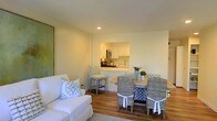 This single-floor space offers a thoughtfully arranged open-concept layout that seamlessly connects the living, dining, and kitchen areas. The interior is finished with warm hardwood-style flooring throughout, contributing to a cohesive and inviting atmosphere. Neutral, cream-toned walls reflect natural and recessed lighting, keeping the space feeling bright and airy.

The living area features a classic white slipcovered sofa accented with patterned throw pillows in soft blue and white tones, establishing a relaxed, coastal-inspired design theme. Adjacent to the living space, the dining area accommodates a wooden table surrounded by woven rattan chairs with coordinating blue and white tie-back cushions, reinforcing the casual yet refined aesthetic.

A pass-through window connects the dining area directly to the kitchen, enhancing the open feel of the layout. The kitchen is outfitted with white cabinetry, stainless steel appliances including a microwave and range, and a countertop that extends to form a convenient breakfast bar with patterned bar seating. The overall ambiance is light, clean, and welcoming, with a consistent design palette carried throughout.