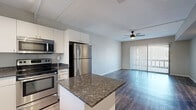 This single-floor residence showcases a thoughtfully designed open-concept layout that seamlessly connects the kitchen and living areas. The kitchen is well-appointed with a full suite of stainless steel appliances, including a refrigerator, range, built-in microwave, and dishwasher. Crisp white cabinetry extends to the ceiling, offering generous storage space, while dark granite countertops provide a striking contrast. A peninsula with matching granite extends the counter space and serves as a natural divider between the kitchen and the adjacent living area.

The living space is finished with wood-style flooring in a cool gray tone that runs throughout, lending a modern, cohesive aesthetic. A ceiling fan is centrally positioned overhead, adding both function and a finishing touch to the space. A large sliding glass door fitted with vertical blinds dominates one wall, allowing natural light to flood the interior while providing access to an outdoor area.

A dedicated entry corridor connects the front door to the main living space, flanked by neutral gray walls and consistent flooring. The overall color palette—neutral grays, whites, and warm wood tones—creates a clean, contemporary ambiance throughout the residence.