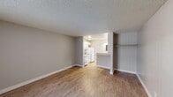 Located at 629 S Main St in Las Vegas, NV, this single-floor unit offers a practical and efficiently designed layout. The open living area is finished with warm wood-look laminate flooring throughout, neutral gray walls, and a textured popcorn ceiling that adds a classic residential character. A window along the front wall allows natural light to fill the space, complemented by a wall-mounted AC unit for climate control. The entry door opens directly into this main living space, creating a straightforward and accessible flow.

Toward the rear, the kitchen is partially separated by a half-wall that doubles as a breakfast bar with a laminate countertop — providing a functional divide while maintaining an open feel. The kitchen is equipped with white cabinetry, a white refrigerator, a gas range with oven, and a single-basin sink, all set against matching wood-look flooring. The cabinetry offers practical overhead storage, and the layout is compact yet well-organized. Adjacent to the kitchen, a bathroom is visible with a mirrored vanity and wall lighting. A built-in open closet with a hanging rod is also present, offering convenient storage within the main living space. The overall ambiance is clean, neutral, and move-in ready.