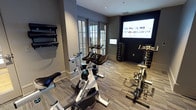 This single-floor fitness space is thoughtfully designed to accommodate a range of exercise needs in a compact yet efficient layout. The room features multiple Spinning bikes positioned as a focal point, making it well-suited for cycling-based workouts. A large wall-mounted display screen faces the bikes, enabling users to follow guided workout sessions or fitness content during their training.

The flooring consists of durable, patterned carpet tiles in neutral tones, providing both comfort underfoot and a polished aesthetic. The walls are finished in a deep charcoal gray, lending the space a modern, focused atmosphere conducive to exercise. Natural light enters through French-style glass doors along one wall, offering a connection to the outdoors while maintaining an energetic interior ambiance.

Along the perimeter, a structured dumbbell rack holds a full range of free weights, supported by additional weight storage racks, ensuring that strength training needs are well met. Wall-mounted equipment storage is also visible, adding to the room's organizational efficiency. Recessed lighting supplements the natural light, keeping the space bright and functional throughout the day.