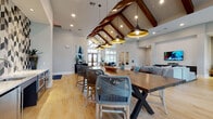 This single-floor community clubhouse is a thoughtfully designed gathering space that combines architectural elegance with contemporary comfort. The centerpiece of the space is a dramatic vaulted ceiling adorned with rich, dark-stained wood beams, complemented by pendant lighting with gold-toned interiors that cast a warm glow over the open-concept layout. Light hardwood flooring runs throughout, enhancing the bright and airy atmosphere.

The main gathering area features generously sized communal tables with woven rope-style chairs, ideal for socializing or collaborative use. Tall floor-to-ceiling windows and glass entry doors flood the space with natural light and offer views of the surrounding exterior. A sculptural live-edge wood console table serves as a striking focal point near the entrance, paired with a tufted bench seat.

The lounge area is equipped with comfortable seating arrangements centered around a wall-mounted television set into a shiplap accent wall, creating a relaxed retreat atmosphere. A dedicated nook showcases a curated gallery wall of framed artwork alongside a geometric-patterned area rug, adding an artistic dimension to the space. The entry corridor features a bold chevron-patterned tile accent wall and beverage appliances, offering a welcoming and functional arrival experience.