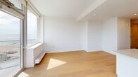 Located at 1 Ocean Dr in Brooklyn's Coney Island neighborhood, this modern one-bedroom, one-bathroom residence offers a thoughtfully designed layout across a single floor. Light hardwood flooring runs throughout, complementing the clean, white interiors that define the space's contemporary aesthetic.

The open-plan kitchen and living area serves as the heart of the home. The kitchen features sleek, high-gloss cabinetry, stainless steel appliances including a refrigerator, microwave, and range, and a peninsula with a quartz countertop that doubles as a breakfast bar. Pendant lighting above the peninsula adds a refined touch. Floor-to-ceiling windows and a glass-panel balcony door flood the living area with natural light and frame expansive views of the Brooklyn skyline and beyond — a standout feature of this residence.

The bedroom is a well-proportioned retreat with light wood flooring, a large closet with bi-fold doors, and a window offering additional city views. The bathroom is finished with large-format stone-look floor tiles, marble-accented tub surround, a soaking tub with shower, and a warm wood vanity with an integrated sink.

Additional highlights include a private balcony, in-unit laundry, a hallway with two closets, and recessed lighting throughout.