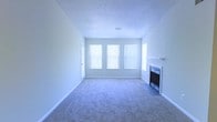 This single-floor space presents a straightforward and functional layout centered around a spacious living area. The room is finished with wall-to-wall carpeting in a neutral tone, contributing to a clean and understated aesthetic throughout. A textured ceiling adds subtle dimension overhead, while white baseboards and light gray walls create a bright, cohesive color palette.

A standout feature of the living area is the fireplace positioned along one wall, framed by a simple white mantel and fitted with a glass-front insert, offering both a functional heating element and a focal point for the room. Adjacent to the fireplace, a side window provides additional light from a secondary angle.

Natural light is a notable characteristic of this space, with three large windows along the main wall fitted with vertical blinds that allow for adjustable light control. The windows contribute to an open and airy feel, brightening the interior throughout the day. A single interior door provides access to additional areas of the unit. Overall, the space offers a clean, move-in-ready environment with neutral finishes suited to a variety of interior styles.