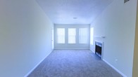 This single-floor space presents a straightforward and functional layout centered around a spacious living area. The room is finished with wall-to-wall carpeting in a neutral tone, contributing to a clean and understated aesthetic throughout. A textured ceiling adds subtle dimension overhead, while white baseboards and light gray walls create a bright, cohesive color palette.

A standout feature of the living area is the fireplace positioned along one wall, framed by a simple white mantel and fitted with a glass-front insert, offering both a functional heating element and a focal point for the room. Adjacent to the fireplace, a side window provides additional light from a secondary angle.

Natural light is a notable characteristic of this space, with three large windows along the main wall fitted with vertical blinds that allow for adjustable light control. The windows contribute to an open and airy feel, brightening the interior throughout the day. A single interior door provides access to additional areas of the unit. Overall, the space offers a clean, move-in-ready environment with neutral finishes suited to a variety of interior styles.