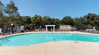 This outdoor community amenity area is centered around a generously sized swimming pool with clearly marked depth gradients, offering a clean and inviting aquatic space for recreation and relaxation. The pool's vivid turquoise water is complemented by a wide concrete sun deck that lines the perimeter, furnished with numerous lounge chairs ideal for sunbathing. A white pergola structure serves as a focal point along the poolside, providing a shaded gathering spot between the deck and the lush tree line that borders the property, creating a natural privacy screen. Adjacent to the pool, a wooden covered pavilion with picnic tables offers additional shaded seating for socializing or outdoor dining. The pool house building, clad in weathered cedar shake siding and accented with classic lantern-style lighting, houses a unisex restroom facility for convenience. Posted pool rules, safety signage, and an emergency contact board are prominently displayed on the building's exterior, reflecting a commitment to safety. A tennis court is also visible in the background, adding to the range of outdoor recreational options available within this community amenity space. The entire area is enclosed by a perimeter fence, ensuring a secure and well-maintained environment.