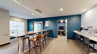 Located at 201 W Glenn Ave in Auburn, Alabama, this well-appointed amenity space offers a thoughtfully designed single-floor layout combining social and professional functionality. The lounge area is a design standout, featuring a bold geometric black-and-white accent wall, modern seating arrangements with gray sofas and brown leather chairs accented by vibrant yellow throw pillows, and a glass coffee table. Linear recessed lighting and large windows allow natural light to fill the space while offering views of the surrounding exterior. Patterned carpet tiles with subtle yellow detailing add texture to the floor throughout. The conference room is equipped for productivity, centered around a long wood-grain conference table surrounded by ergonomic mesh chairs. A wall-mounted flat-screen display and a large glass whiteboard provide versatile presentation options, while a deep blue textured accent wall adds a professional yet inviting atmosphere. A triptych of colorful abstract artwork adorns one wall, lending a creative touch. Additional workspace is available through a bar-height counter seating area along one wall of the lounge, ideal for individual work or casual collaboration. Windows throughout the space provide consistent natural light and views of the neighboring courtyard and buildings, creating a bright and connected environment.