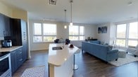 This well-appointed 2-bedroom, 2-bathroom residence offers a thoughtfully designed open-concept floor plan that seamlessly connects the kitchen, dining, and living areas into one cohesive and inviting space. The kitchen serves as the centerpiece, featuring a large island with a striking white quartz countertop, an undermount sink, and stylish pendant lighting that adds a modern touch. Dark hardwood-style flooring runs throughout the main living area, providing a warm contrast to the bright, neutral-toned walls. Multiple large windows wrap around the living and dining areas, flooding the space with natural light and offering expansive outdoor views that enhance the airy atmosphere. The living area provides generous room for comfortable seating arrangements, while the open dining space offers a natural gathering area adjacent to the kitchen island. Track lighting overhead supplements the natural light, ensuring the space remains well-lit at any time of day. The overall ambiance is clean, contemporary, and welcoming, with a layout that maximizes both functionality and flow — ideal for everyday living and entertaining alike.