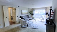 This single-floor residence showcases a thoughtfully designed living room that serves as the focal point of the space. Large sliding glass doors span one wall, flooding the interior with abundant natural light and offering views of lush greenery outside, creating a seamless connection between indoors and outdoors. The living room is finished with plush wall-to-wall carpeting in a warm neutral tone, contributing to a comfortable and inviting atmosphere. A coordinated seating arrangement, featuring a grey sofa and two upholstered accent chairs with nailhead trim, is anchored by a patterned area rug, adding visual depth and texture to the space. A glass-topped coffee table serves as a functional centerpiece. A large, colorful canvas artwork adorns one wall, complemented by a tall decorative plant that introduces an organic element. Neutral-toned walls and a smooth ceiling with recessed HVAC vents enhance the clean, understated aesthetic. A doorway visible at the left provides access to adjacent rooms, suggesting a practical and well-connected floor plan throughout the residence.