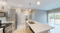Located at 3800 E Campus Ave in Holland, Michigan, this one-bedroom, one-bathroom unit offers a thoughtfully designed single-floor layout with modern finishes throughout. The open-concept kitchen and living area serves as the heart of the unit, featuring light wood-look flooring that enhances the bright, airy feel of the space. The kitchen is equipped with stainless steel appliances — including a refrigerator, range, and microwave — complemented by sleek, taupe cabinetry with matte black hardware and a quartz-topped island with a double basin sink. Pendant lighting above the island adds a contemporary touch. The living area benefits from a sliding glass door that invites natural light and provides access to an outdoor space. The bedroom is carpeted for comfort and includes a closet with built-in shelving, as well as a window with outdoor views, creating a calm and private retreat. The bathroom is clean and functional, outfitted with a vanity with under-sink storage, a mirror with overhead lighting, and a shower-tub combination. Neutral warm-toned walls and white trim carry consistently throughout, giving the unit a cohesive and modern aesthetic.