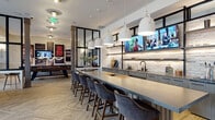 This single-floor communal lounge is thoughtfully designed to serve as a multifunctional social hub. The space is anchored by a long bar-height counter topped with a sleek grey surface and lined with upholstered velvet bar stools, positioned in front of a textured stacked-stone accent wall. Above the counter, pendant lights with large white bell shades provide focused illumination, while wall-mounted flat-screen TVs and open shelving with under-shelf lighting add both function and visual interest. The bar area features built-in cabinetry, a utility sink, and draft beer taps, making it well-equipped for entertaining.

The open lounge area features eye-catching herringbone-patterned wood-look flooring that runs throughout, lending warmth and texture underfoot. Structural columns punctuate the space, complementing the industrial aesthetic. Tufted cognac leather banquette seating is positioned along floor-to-ceiling windows, offering street views and abundant natural light, accompanied by white-topped tables with gold pedestals.

A dedicated nook serves as a quieter meeting area, enclosed with deep teal paneled walls, a round black table with upholstered chairs, a mounted TV, and large windows overlooking a residential streetscape. Toward the rear, a pool table adds a recreational element, completing this versatile, well-appointed communal space.