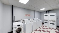 This single-floor shared laundry facility is designed with practicality and functionality in mind. The room features a row of top-loading washing machines aligned along one wall, accompanied by a front-loading washer, providing users with multiple options for their laundry needs. To the right, stacked dryer units maximize the use of vertical space, ensuring a comprehensive laundry solution within a compact footprint.

The space is finished with a classic red and white checkered tile floor, adding a utilitarian yet distinct visual character to the room. Overhead, a drop ceiling with recessed fluorescent panel lighting ensures the area is well-lit throughout. The walls are painted in a neutral light gray, contributing to a clean and orderly atmosphere.

Informational signage is posted on the walls, providing users with guidelines for proper use and upkeep of the shared facility. The overall layout is straightforward and efficient, with machines arranged to allow easy access and circulation. This laundry room serves as a well-equipped communal amenity, maintained to support the everyday needs of its users.