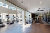 This single-floor fitness center is thoughtfully designed to accommodate a full range of exercise disciplines within a bright, open layout. The space is finished with rich dark hardwood flooring, crisp white crown molding, and soft sage-green accent walls, creating a polished yet energizing atmosphere. Recessed lighting and multiple modern black ceiling fans are distributed throughout, ensuring consistent airflow and illumination across all zones.

The cardio area features a row of treadmills and elliptical machines positioned along floor-to-ceiling windows, allowing exercisers to enjoy expansive outdoor views and generous natural light that floods the space. Adjacent to this is a dedicated strength training zone, equipped with a full dumbbell rack, cable machines, weight benches, and free-weight stations arranged on rubber-matted flooring for safety and stability.

A separate cycling studio, enclosed by sleek glass partition walls, houses several stationary spin bikes and is outfitted with wall-mounted flat-screen televisions and bold artwork, creating an immersive environment for group or solo cycling sessions. A small seating area with a bistro-style table and chairs provides a convenient space to rest or review workout plans. The overall design balances functionality with aesthetic refinement, making this fitness center both highly practical and visually welcoming.