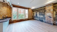 Located at 266 E Michigan Ave in the heart of downtown Kalamazoo, this 2-bedroom, 1-bathroom unit offers a compelling blend of historic character and contemporary design. Light hardwood flooring flows throughout the single-floor layout, complementing the clean white walls and modern finishes found across every room. The open living, dining, and kitchen area serves as the centerpiece of the unit. The kitchen is well-appointed with white shaker-style cabinetry, dark granite countertops, a subway tile backsplash, and stainless steel appliances including a range, microwave, and refrigerator, with a large window framing views of the surrounding downtown architecture. The living area features striking exposed brick walls adorned with original painted lettering — a nod to the building's industrial heritage — alongside large windows that invite abundant natural light. Both bedrooms offer comfortable proportions, with one showcasing an eye-catching exposed brick accent wall with historic signage. The hallway features an exposed metal duct, adding to the loft-inspired aesthetic. The bathroom includes a full tub-shower combination with white tile surround and matte black fixtures. A dedicated closet and hallway complete this thoughtfully designed urban residence.