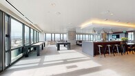 Located in Denver's Golden Triangle neighborhood, the Pool Lobby at 1140 Bannock St is a generously sized amenity space designed with a modern, sophisticated aesthetic. Floor-to-ceiling windows wrap the perimeter, flooding the interior with abundant natural light and framing panoramic views of the Denver skyline and the Rocky Mountains beyond.

The open-plan layout is divided into distinct zones, each thoughtfully appointed. A game area features a full-size billiards table and a shuffleboard table set atop large-format light stone tile flooring, creating a clean and airy atmosphere. Adjacent to this is a stylish bar and kitchen station with a long island countertop, bar seating, pendant lighting, and rich wood cabinetry equipped with professional appliances—ideal for entertaining.

A lounge and social area transitions the space into a warmer, more intimate setting, anchored by a linear gas fireplace set within a stone surround. A rich wood-paneled ceiling overhead adds warmth, while a flat-screen TV, built-in shelving, and curated artwork contribute to the refined atmosphere. Seating throughout includes contemporary sofas, lounge chairs, and café-style tables arranged atop a vivid blue area rug.

Additional window-side café seating areas invite relaxation with unobstructed city views, rounding out this well-equipped, design-forward communal space.