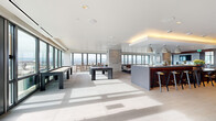Located in Denver's Golden Triangle neighborhood, the Pool Lobby at 1140 Bannock St is a generously sized amenity space designed with a modern, sophisticated aesthetic. Floor-to-ceiling windows wrap the perimeter, flooding the interior with abundant natural light and framing panoramic views of the Denver skyline and the Rocky Mountains beyond.

The open-plan layout is divided into distinct zones, each thoughtfully appointed. A game area features a full-size billiards table and a shuffleboard table set atop large-format light stone tile flooring, creating a clean and airy atmosphere. Adjacent to this is a stylish bar and kitchen station with a long island countertop, bar seating, pendant lighting, and rich wood cabinetry equipped with professional appliances—ideal for entertaining.

A lounge and social area transitions the space into a warmer, more intimate setting, anchored by a linear gas fireplace set within a stone surround. A rich wood-paneled ceiling overhead adds warmth, while a flat-screen TV, built-in shelving, and curated artwork contribute to the refined atmosphere. Seating throughout includes contemporary sofas, lounge chairs, and café-style tables arranged atop a vivid blue area rug.

Additional window-side café seating areas invite relaxation with unobstructed city views, rounding out this well-equipped, design-forward communal space.
