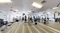Located at 228 Dixie Dr in Tallahassee, FL, this fitness center offers a comprehensive workout facility within a single, open-plan floor. The space is designed to maximize functionality, featuring striped low-pile carpeting throughout that defines the workout areas while providing comfort underfoot. Multiple ceiling fans are suspended from the textured white ceiling, ensuring consistent airflow and a comfortable exercise environment.

The facility is well-lit by a combination of fluorescent overhead fixtures and natural light streaming in through several windows fitted with blinds, creating a bright and energizing atmosphere. A full-length wall of mirrors along one side visually expands the space and allows users to monitor their form during workouts.

The strength training area is outfitted with Cybex cable machines, a power rack with a flat bench, a comprehensive dumbbell rack with free weights, and a variety of resistance machines. The cardio section includes multiple treadmills, stair climbers, and an elliptical, catering to a wide range of fitness routines. Stability balls and additional accessories are available for functional training. A wall-mounted flat-screen television adds an element of entertainment. Direct exterior door access provides convenient entry and exit to the facility.