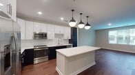 Located at 600 Willard St in Durham, NC, this single-floor residence offers a thoughtfully designed open-concept layout that seamlessly connects the kitchen and living areas. Rich, dark hardwood-style flooring flows throughout the main living spaces, providing a warm and contemporary foundation.

The kitchen is a standout feature, showcasing a bold two-tone design with sleek dark lower cabinets and crisp white upper cabinetry, all accented by brushed nickel hardware. A white quartz countertop extends across the workspace and onto a generous central island, ideal for casual dining or meal prep. White subway tile backsplash, stainless steel appliances, and a set of three industrial-style pendant lights complete this well-appointed culinary space.

The open living area benefits from large windows fitted with blinds, allowing ample natural light to fill the room. A glass door provides access to an outdoor area with views of the surrounding neighborhood.

The bathroom is finished in a clean, all-white palette featuring subway tile surround, a soaking tub, and a vanity with brushed nickel fixtures. Light tile flooring enhances the bright, crisp feel of the space. A spacious walk-in closet with multi-tiered wire shelving and soft carpet underfoot offers generous storage and organizational flexibility throughout.