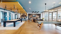 Located at 2554 Davis Rock Dr in Dacula, Georgia, this single-floor amenity space is a thoughtfully designed communal retreat that blends contemporary style with inviting warmth. The living room serves as the centerpiece, anchored by a dramatic exposed brick accent wall fitted with built-in open shelving and a wall-mounted flat-screen television. A continuous built-in bench seat with colorful throw pillows lines the base of the wall, offering casual seating. The ceiling is painted a bold teal-blue, complemented by a cluster of black-and-gold dome pendant lights that add a striking visual focal point overhead. Light hardwood flooring in a chevron herringbone pattern flows throughout, adding texture and elegance underfoot. The space is furnished with plush, curved lounge chairs, a dark wood communal table with leather bench seating, and a full-size pool table positioned beneath a floating wood-panel ceiling feature with sleek cylindrical pendant lights. Large floor-to-ceiling glass doors and windows allow abundant natural light to pour in, offering views of the outdoor patio area. The hallway entry is bright and airy, featuring decorative artwork and a seamless transition into the main lounge. The overall ambiance is modern, vibrant, and welcoming — ideal for socializing and recreation.