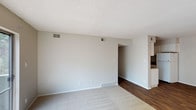 This single-floor residence presents a straightforward and functional layout designed for comfortable everyday living. The main living area is finished with soft, neutral-toned carpeting that creates a warm and understated atmosphere. Walls are painted in a light gray hue, contributing to a bright and airy feel throughout the space. A textured popcorn ceiling adds a classic touch overhead, while multiple ceiling-mounted HVAC vents ensure consistent climate control throughout the living area. An open archway connects the carpeted living space to an adjoining area finished with rich, dark hardwood-style flooring, offering a natural transition between zones. This secondary area leads further into the residence, hinting at additional rooms beyond. Baseboard heating elements line the lower walls, supplementing the overhead ventilation system. The space benefits from clean white trim and baseboards that frame the rooms neatly. The overall design is neutral and move-in ready, providing a blank canvas well-suited for a variety of interior styles and furnishing preferences. The layout flows openly, making the space feel larger than its footprint, and the consistent color palette ties each area together cohesively.