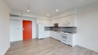 This single-floor space presents a clean, contemporary design with a bright and open layout. The kitchen is the focal point of the unit, outfitted with sleek flat-front white cabinetry that extends to the upper walls, maximizing storage throughout. White appliances — including a full-size refrigerator, range, microwave, and dishwasher — complement the crisp, minimalist aesthetic. A mosaic tile backsplash in muted gray tones adds a subtle decorative texture behind the countertops, contrasting tastefully against the white surfaces. Recessed lighting installed in the white ceiling provides clean, even illumination across the space. The light-toned laminate flooring runs throughout, contributing to an airy, cohesive feel underfoot. A bold red entry door serves as a striking architectural accent, introducing a pop of color that enlivens the otherwise neutral palette. The open configuration of the kitchen and living area creates a sense of spaciousness, allowing for flexible furniture arrangement. Overall, the space strikes a balance between modern functionality and understated style, offering a well-appointed interior that is both practical and visually inviting.