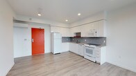 This single-floor space presents a clean, contemporary design with a bright and open layout. The kitchen is the focal point of the unit, outfitted with sleek flat-front white cabinetry that extends to the upper walls, maximizing storage throughout. White appliances — including a full-size refrigerator, range, microwave, and dishwasher — complement the crisp, minimalist aesthetic. A mosaic tile backsplash in muted gray tones adds a subtle decorative texture behind the countertops, contrasting tastefully against the white surfaces. Recessed lighting installed in the white ceiling provides clean, even illumination across the space. The light-toned laminate flooring runs throughout, contributing to an airy, cohesive feel underfoot. A bold red entry door serves as a striking architectural accent, introducing a pop of color that enlivens the otherwise neutral palette. The open configuration of the kitchen and living area creates a sense of spaciousness, allowing for flexible furniture arrangement. Overall, the space strikes a balance between modern functionality and understated style, offering a well-appointed interior that is both practical and visually inviting.