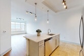 Located at 25 W Tremont Ave in Charlotte's vibrant South End neighborhood, this well-appointed 1-bedroom, 1-bathroom residence offers a thoughtfully designed open-concept layout. The combined kitchen and living area is anchored by a generous center island with a quartz countertop, undermount sink, and stainless steel dishwasher, all illuminated by elegant glass pendant lights and a track lighting system. The kitchen features white shaker cabinetry, a herringbone-patterned tile backsplash, and a full suite of stainless steel appliances, including a French door refrigerator and range. Light-toned stone tile flooring grounds the kitchen, while the living area transitions to warm light hardwood floors, filling the space with a bright, airy feel. Large floor-to-ceiling windows flood the living room with natural light and frame sweeping views of the Charlotte skyline and surrounding cityscape. The bedroom is finished with plush carpeting and its own oversized window offering panoramic views. The bathroom is elegantly appointed with travertine tile flooring, a soaking tub set within an arched alcove with coordinating tile surround, and a white vanity with quartz countertop — creating a refined, spa-like retreat.