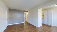 This single-floor residence offers a straightforward and functional layout designed for comfortable everyday living. The main living area is finished with warm, wood-style flooring that runs throughout, complementing the crisp white trim and neutral-toned walls — including a tasteful gray accent wall that adds subtle depth to the space. Recessed HVAC venting is integrated into the ceiling, contributing to a clean, uncluttered look overhead. A partial wall divider separates the living area from the kitchen, creating a sense of openness while maintaining distinct zones. The kitchen features white cabinetry, light stone-toned countertops, and white appliances, lending a bright and tidy aesthetic. A hallway visible from the main living area leads further into the residence, suggesting additional rooms beyond. Natural light filters in through a window, enhancing the airy feel of the space. The overall ambiance is clean, minimal, and move-in ready, with a neutral palette that provides a versatile backdrop suitable for a variety of interior styles and furnishings.