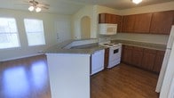 This single-floor home offers a well-designed, open-concept layout that seamlessly connects the kitchen and living areas. The kitchen is a standout feature, showcasing warm-toned wood cabinetry that provides ample storage alongside granite-style countertops that add a touch of elegance. A central peninsula extends the counter space, making it ideal for meal preparation or casual dining. White appliances, including a range, dishwasher, and over-the-range microwave, are neatly integrated into the layout for a clean, cohesive look. Rich hardwood flooring runs throughout the space, lending warmth and continuity to the interior. An arched entryway adds an architectural detail that enhances the charm of the open floor plan. Natural light filters in through a window near the entrance, brightening the living area and creating a welcoming atmosphere. A ceiling fan is also present, contributing to the comfort of the space. The overall ambiance is practical yet inviting, with a layout that balances functionality and livability in a compact, efficient single-level design.