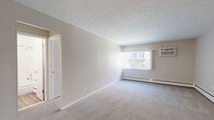 Located at 19740 Lorain Road in Fairview Park, Ohio, this one-bedroom, one-bathroom unit offers a practical and comfortable single-floor layout. The living room is the largest area of the unit, featuring neutral-toned carpet, a textured ceiling, and a large window that allows natural light to fill the space. Baseboard heating runs along the walls, and a wall-mounted air conditioning unit provides climate control year-round. The open feel of the living room is enhanced by clean, light-colored walls and generous floor space suitable for a variety of furniture arrangements. A ceiling fan adds additional comfort and airflow to the common area. The bedroom is a well-proportioned private retreat, also carpeted, with its own window, baseboard heating, and a full-width bifold closet offering ample storage. The bathroom features classic white square tile surrounding a full-length soaking tub with an overhead shower, complemented by a coordinating toilet and a countertop with a speckled finish. A separate area near the bedroom provides additional closet or storage space. The unit's neutral palette and functional layout make it a versatile and move-in-ready space.