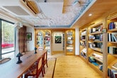 This single-floor commercial space offers a well-appointed and cohesive interior defined by warm natural wood flooring, exposed ceiling infrastructure, and a carefully curated mix of functional zones. The main lounge area serves as the heart of the space, featuring built-in bench seating upholstered in textured, patterned fabric, complemented by a small pedestal table and layered area rugs. Floor-to-ceiling glazed windows flood this area with abundant natural light while framing direct views of the surrounding streetscape and tree-lined neighborhood. A decorative string curtain partition adds visual interest and subtly separates zones without enclosing them. The library-style reading and meeting room is lined with illuminated built-in wood shelving stocked with books and curated objects, creating an intimate yet intellectual atmosphere. A solid wood dining-style table with cane-back chairs anchors this room, making it equally suitable for focused work or group gatherings. Exposed brick columns and textured wall surfaces add an industrial warmth throughout. A private alcove provides a quiet retreat with a stone-topped desk, fabric-lined walls, and a narrow vertical window. The overall ambiance is eclectic yet refined, blending residential comfort with a functional, creative commercial sensibility.