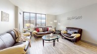 This thoughtfully designed 1-bedroom, 1-bathroom model unit is spread across a single floor, offering a cohesive and functional layout ideal for comfortable urban living. The living room serves as the heart of the home, featuring light wood-style flooring and expansive floor-to-ceiling windows that flood the space with natural light and frame sweeping city skyline views. The open feel of this room makes it ideal for both relaxing and entertaining. The galley-style kitchen is efficiently laid out with warm wood cabinetry, light countertops, white appliances including a gas range and refrigerator, and clean tile flooring that maximizes the available space. The bedroom is generously proportioned with soft wall-to-wall carpeting, neutral tones, and a large window offering additional city views, creating a calm and restful retreat. A second bedroom configuration is also visible in the model, providing flexible living options. The bathroom is clean and functional, featuring tile flooring, a vanity with storage, and a tub-shower combination. Throughout the unit, the neutral color palette and abundant natural light contribute to a bright, airy ambiance that feels both welcoming and versatile.