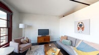 This single-floor space showcases a compelling blend of industrial heritage and contemporary design. Throughout the living areas, whitewashed exposed brick walls serve as a defining architectural feature, adding texture and warmth while maintaining a bright, airy atmosphere. Dark hardwood-style flooring grounds the space and contrasts effectively with the light-toned walls and ceilings.

The living room is thoughtfully arranged with mid-century modern furnishings, including a tufted sofa, accent chair, and a walnut media credenza, all anchored by a patterned area rug. Large, arched windows with deep crimson frames flood the space with natural light and offer elevated views of the surrounding neighborhood, creating a dramatic focal point.

A dedicated sitting nook near the windows features a vibrant accent chair and a marble-topped bistro table, ideal for casual use. The bedroom continues the whitewashed brick aesthetic, accented by a warm pendant light and a gold-framed vanity with a round mirror.

The kitchen is equipped with stainless steel appliances, white cabinetry, a subway tile backsplash, and an exposed industrial duct overhead, reinforcing the loft-inspired character. The bathroom features a streamlined white vanity with a large mirror, neutral tile flooring, and a shower-tub combination.