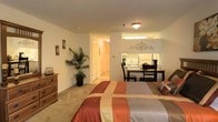 This single-floor unit features an open-concept layout that thoughtfully integrates the sleeping, dining, and kitchen areas into one functional space. The main living area is carpeted and accommodates a full-sized bed dressed in warm earth tones, complemented by a classic wooden dresser along the wall. A potted palm plant adds a natural, organic touch to the corner of the room.

Adjacent to the sleeping area, a compact dining setup with a dark wooden table and chairs sits alongside a partially visible kitchen. The kitchen area features white cabinetry and appliances, illuminated by under-cabinet fluorescent lighting that adds brightness to the workspace. Decorative wall art above the kitchen area contributes a personalized charm.

A hallway visible from the main room leads to additional spaces, including what appears to be a closet and bathroom, providing practical storage and utility. The unit is lit by a combination of a central ceiling fixture and natural ambient light, creating a warm, inviting atmosphere throughout. The neutral wall tones and carpeted flooring lend the space a comfortable, understated aesthetic.