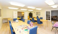 Located at 32 Blacks Lane in Springfield, NJ, this single-floor community activity space is designed to foster engagement and social interaction. The open layout accommodates multiple large tables surrounded by cushioned blue chairs, providing ample seating for group activities, games, and casual gatherings. Light wood flooring runs throughout, lending the space a clean, bright aesthetic that is complemented by crown molding along the ceilings and flush-mounted fluorescent lighting overhead.

A well-stocked bookshelf lines one wall, filled with board games, puzzles, and books, making it an ideal spot for recreational activities. A wall-mounted flat-screen television is positioned for easy viewing from the seating areas, alongside sliding glass door access to an outdoor deck with natural light filtering in. Framed artwork decorates the walls, adding warmth and character to the environment.

An adjoining area serves as a small kitchenette, equipped with a full-size refrigerator, microwave, and cabinetry, offering convenient refreshment options. Additional rooms connect seamlessly, providing flexible space for a variety of programming needs. The overall ambiance is welcoming and functional, thoughtfully arranged to support both structured activities and relaxed social interaction throughout the day.