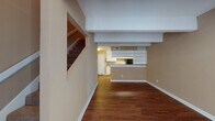 This two-story space is designed with a practical, open layout that connects the main living areas on the ground floor. The lower level features warm hardwood flooring throughout, neutral beige walls, and white trim, creating a cohesive and understated aesthetic. A carpeted staircase with white framing leads to the upper floor, while structural beams add architectural character to the ceiling above the main living area.

The galley-style kitchen is well-equipped with black appliances, including a refrigerator, electric range with overhead ventilation, and dishwasher. White cabinetry lines both sides of the kitchen, complemented by granite-style countertops and a tile backsplash. A deep double-basin sink adds functionality, while a built-in pantry with multiple shelves provides generous storage.

A half-wall peninsula separates the kitchen from the open living area, which benefits from the flow of natural light. The bathroom features a tiled tub-and-shower combination with an accent border, granite-style vanity countertop, and a mirrored medicine cabinet.

The upper level is carpeted and open, featuring a ceiling fan, pendant lighting, and a windowed overlook that brings in additional natural light, creating a comfortable and versatile secondary living space.