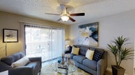 This single-floor residence offers a well-appointed living space designed for both comfort and functionality. The living room serves as the heart of the home, featuring a five-blade ceiling fan with a built-in light fixture that provides both airflow and ambient illumination. A textured ceiling adds a classic touch to the room's character.

Natural light pours in through a large sliding glass door fitted with vertical blinds, offering easy access to a private outdoor patio area where outdoor seating can be enjoyed. The living room comfortably accommodates a plush sofa adorned with decorative pillows in coordinating tones of yellow, navy, and grey, complemented by a sleek glass-top coffee table that adds an open, airy feel to the space.

Bold wall art featuring a dramatic ocean wave composition serves as a striking focal point, lending the room a dynamic yet relaxed aesthetic. The warm, neutral wall tones create a versatile backdrop that complements a variety of décor styles. Overall, the space balances practicality with an inviting ambiance, making it well-suited for everyday living and casual entertaining.