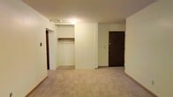 This single-floor space presents a straightforward and functional layout with clean, neutral-toned walls throughout. The main room is finished with wall-to-wall carpeting in a light beige tone, providing a soft underfoot feel and contributing to the overall warmth of the interior. Ceiling-mounted lighting illuminates the space adequately, keeping the atmosphere bright and even.

A prominent open closet is built into the wall, featuring a single hanging rod and a shelf above, offering practical storage without the need for additional furniture. The closet opening is framed cleanly into the wall, maintaining a tidy appearance. Adjacent to the closet, a solid dark wood entry door serves as the main point of access, fitted with a standard lock mechanism. The door's darker tone provides a visual contrast against the light-colored walls.

A secondary doorway is visible, suggesting additional rooms or utility spaces beyond the main area. Baseboards run along the perimeter of the room, adding a modest finishing detail to the overall design. The space is compact yet organized, with a simple, no-frills aesthetic that prioritizes utility and ease of use.