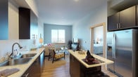 This single-floor residence offers a thoughtfully designed open-concept layout that seamlessly connects the kitchen, dining, and living areas. The kitchen is a standout feature, outfitted with rich dark cabinetry, stainless steel appliances including a range and built-in microwave, and light stone countertops that extend along both sides of the galley-style workspace. A breakfast bar with seating provides a natural transition into the dining and living spaces. Light hardwood-style flooring runs throughout the main living areas, adding warmth to the contemporary palette of soft blue-gray walls. The living room is bright and inviting, anchored by a large window that frames panoramic views of the surrounding cityscape and waterway. The bedroom continues the calming aesthetic with plush carpeting, generous natural light from a wide window overlooking the water, and a walk-in closet for ample storage. A full bathroom features hardwood-style flooring, a soaking tub with shower, dark vanity cabinetry, and stone countertops consistent with the rest of the home. An in-unit laundry closet adds everyday convenience. The overall ambiance is modern and serene, with cohesive finishes and scenic views creating a polished, comfortable living environment.