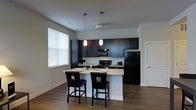 This single-floor residence offers a well-appointed interior with a thoughtfully designed open-concept kitchen that serves as the focal point of the space. The kitchen features rich, dark espresso cabinetry that extends to the upper walls, providing ample storage while creating a striking visual contrast against the light-toned walls. Granite countertops add an elegant touch, complemented by a peninsula breakfast bar with seating for two or more, making it ideal for casual dining or entertaining. Black appliances, including a refrigerator, gas range, and microwave, lend a cohesive and contemporary aesthetic to the cooking space. Two pendant lights hang above the peninsula, providing focused task lighting that enhances the kitchen's modern character. Natural light filters in through a large window fitted with horizontal blinds, brightening the open area. The hardwood-style flooring runs throughout, adding warmth and continuity to the space. The overall ambiance is clean, modern, and functional, with a neutral color palette that allows the dark cabinetry and fixtures to stand out as defining design elements throughout the residence.